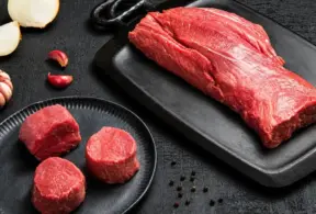 Raw filet mignon steak—lean red meat—on a black cutting board, with three pieces of filet mignon on a black plate, surrounded by garlic, onion, and peppercorns