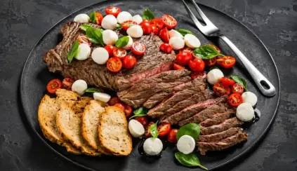 Grilled sliced ​​beef with a cherry tomato and green leaf salad, presented on a dark plate with bread and buffalo mozzarella on the side, illustrating a complete animal protein.