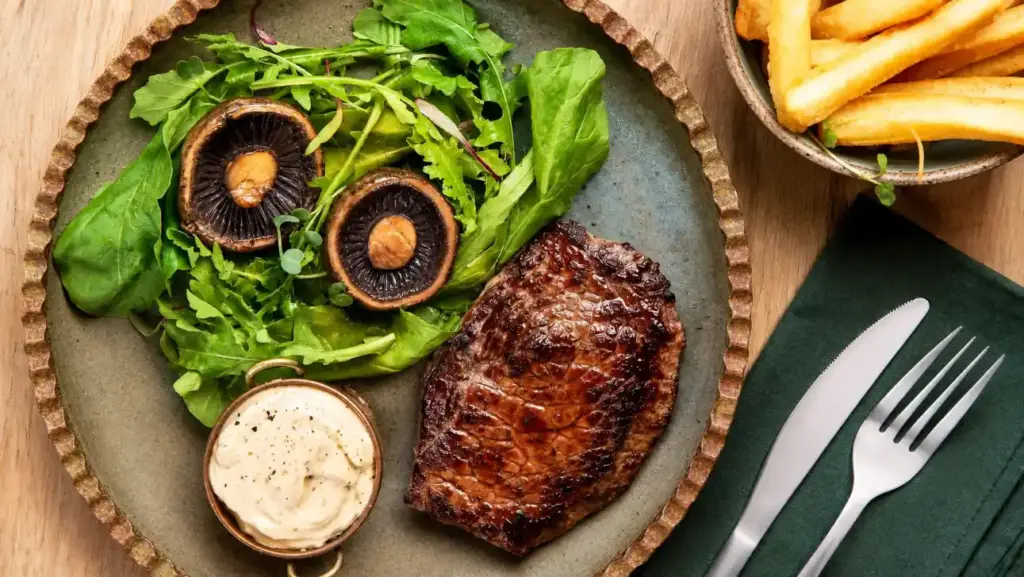 A dish featuring four quick-seared beef cuts—grilled steaks served with a green salad, mushrooms, and a creamy sauce—accompanied by french fries.