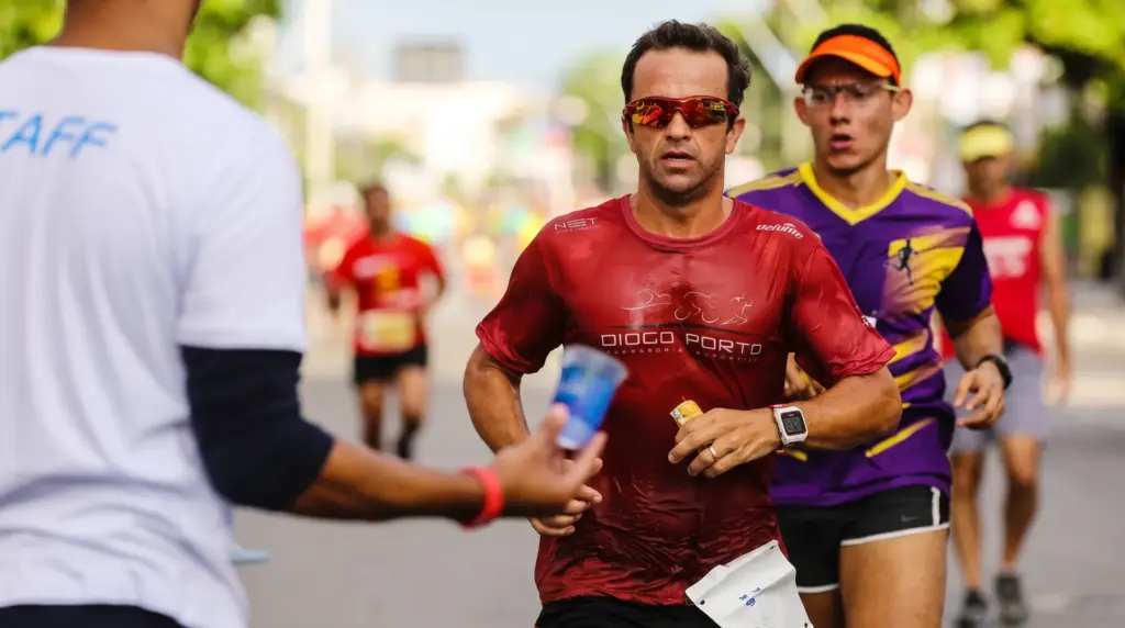 Iron and ferritin: Street marathon—an athlete wearing a red t-shirt and sunglasses runs alongside other participants; the focus is on the runner in the foreground, with a narrator heard in the background during the outdoor race.