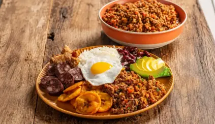 A dish featuring cuts of beef accompanied by a fried egg, beans, and plantain, alongside a bowl of ground meat on a wooden table.