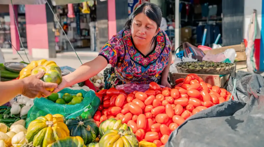 Woman at a food market selecting fresh fruits and vegetables—including pumpkins, bananas, and tomatoes—highlighting food safety.