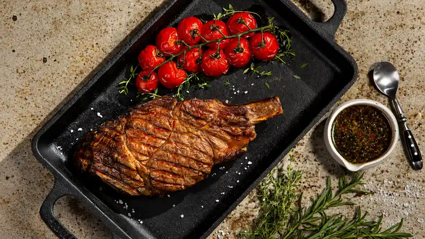 Different meat names: Prime Rib roasted in an iron pan, with herb sprigs, roasted red peppers, and sauce on the side in a small dish. Meat ready to serve.