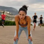Woman running on a coastal road with other people in the background, engaging in outdoor physical activity at sunset in a sporty atmosphere.