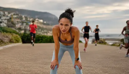 Woman running on a coastal road with other people in the background, engaging in outdoor physical activity at sunset in a sporty atmosphere.