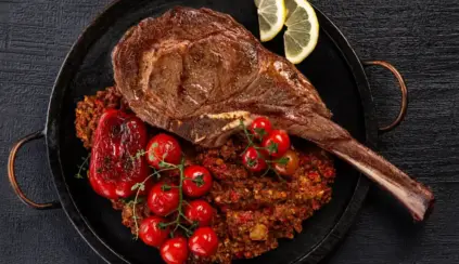 Different meat names: Tomahawk steak pan-seared with crispy-style potatoes, accompanied by cherry tomatoes and lemon slices, set against a dark wood background.
