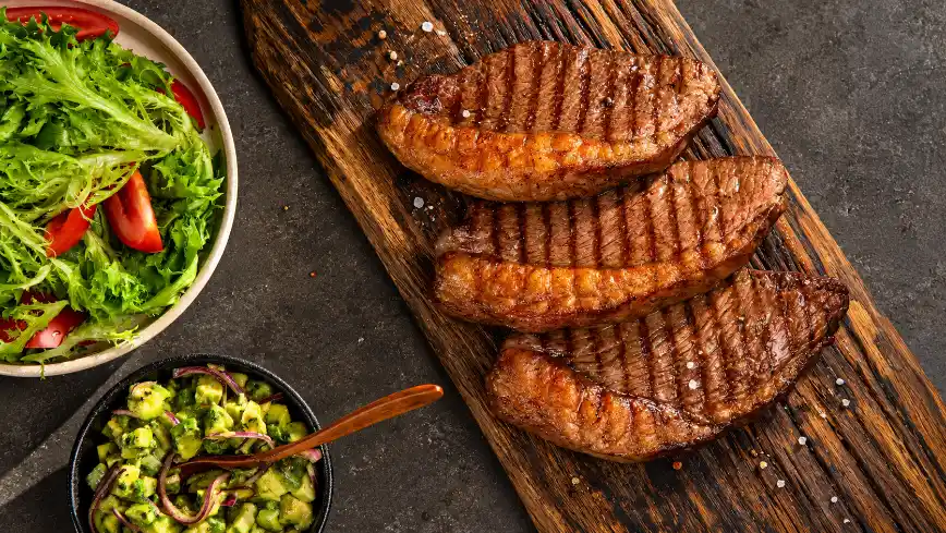 Three grilled steaks on a wooden board sprinkled with coarse salt, alongside a bowl of green salad with tomato slices and a small bowl of chopped avocado salsa on a dark surface—a delicious combination of iron-rich foods, perfect for a healthy and tasty meal.