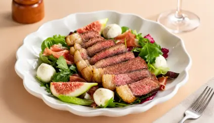 Gourmet salad on a white plate featuring beef slices, mixed greens, arugula, sliced ​​fig, quail eggs, and dressings, set on a light-colored table. Meat science applied to the preparation of dishes using fresh ingredients.