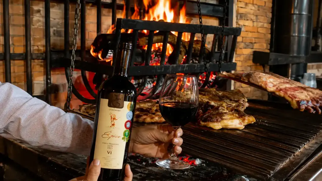 Meat and wine served at a wood-fired grill, featuring a glass and bottle of red wine alongside grilled cuts, in a rustic setting with a fire in the background.