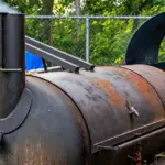 Meat being cooked in a metal barrel grill outdoors; the lid and smoke are visible as the barbecue is being prepared.