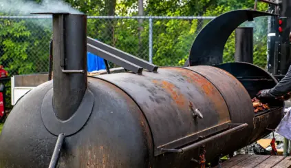 Meat being cooked in a metal barrel grill outdoors; the lid and smoke are visible as the barbecue is being prepared.