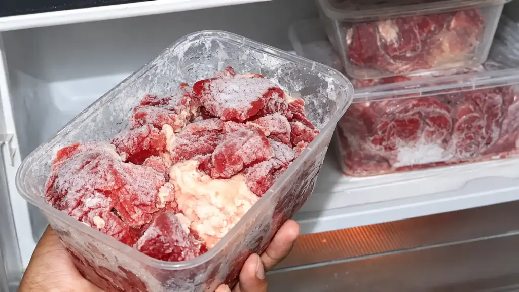 A container of loose meat releasing liquid, containing frozen meat cubes covered in ice, inside a refrigerator next to other containers of meat.