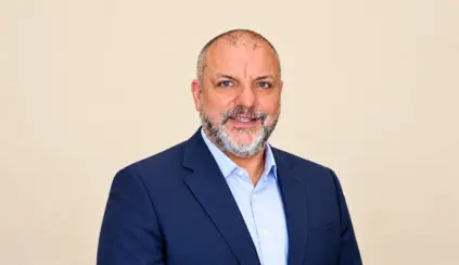 Pedro Loyola wearing a navy blue suit and a light-colored dress shirt, against a plain, light-colored background, smiling at the camera. Corporate headshot.