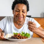 Una señora mayor y de raza negra con expresión feliz almorzando un plato con carne picada, arroz, frijoles y ensalada de lechuga y tomate. Transmitiendo el concepto de la importancia de la carne roja en su edad.