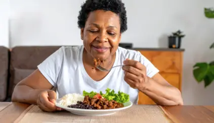 Una señora mayor y de raza negra con expresión feliz almorzando un plato con carne picada, arroz, frijoles y ensalada de lechuga y tomate. Transmitiendo el concepto de la importancia de la carne roja en su edad.