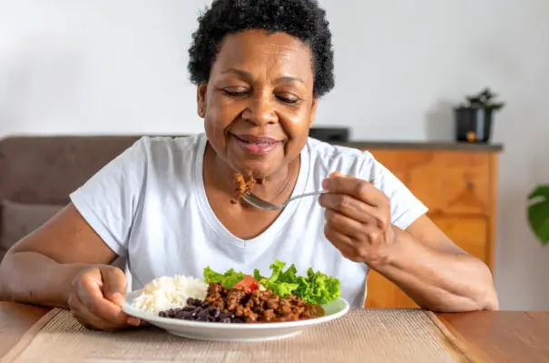Una señora mayor y de raza negra con expresión feliz almorzando un plato con carne picada, arroz, frijoles y ensalada de lechuga y tomate. Transmitiendo el concepto de la importancia de la carne roja en su edad.