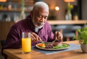 Hombre mayor con cabello blanco, vistiendo una camisa morada, come una comida balanceada con carne de res, frijoles y vegetales, también con un vaso de jugo de naranja al lado del plato, destacando la importancia de la vitamina B2 en la alimentación, la cual el cuerpo no produce de forma natural, pero se encuentra en la carne de res.