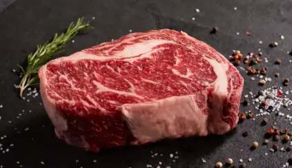 No toda la grasa de la carne es igual: conozca la grasa intramuscular de la carne vacuna, en un corte de carne roja con finas vetas de grasa blanca, bajo especias y sal gruesa sobre una superficie oscura.