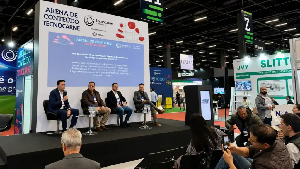 Cinco hombres están sentados en un escenario frente a una pantalla grande durante una conferencia, dirigiéndose a la audiencia. Al fondo, se aprecian stands de exposición y señalización del evento TecnoCarne y del Content Arena, lo que resalta la importancia de TecnoCarne —y de la cadena de suministro cárnico— dentro del sector