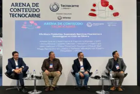 Cuatro hombres vestidos con ropa de negocios están sentados en el escenario, frente a un telón de fondo azul y blanco, en TecnoCarne. Parecen formar parte de un panel de discusión sobre cómo superar las barreras de productividad en las fábricas de la cadena de suministro cárnico