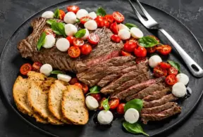 Carne vacuna a la parrilla con láminas y ensalada de tomates cherry y hojas verdes, presentada en un plato oscuro con pan y mozzarella de búfala a un lado, ilustrando una proteína animal completa.