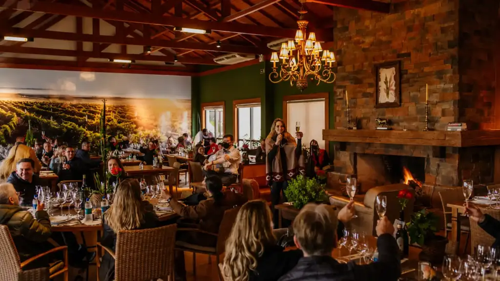 Carne y vino en una celebración: personas brindando en un comedor rústico con la mesa servida, copas de vino y un ambiente iluminado por lámparas de araña, junto a una chimenea y una pared de piedra.
