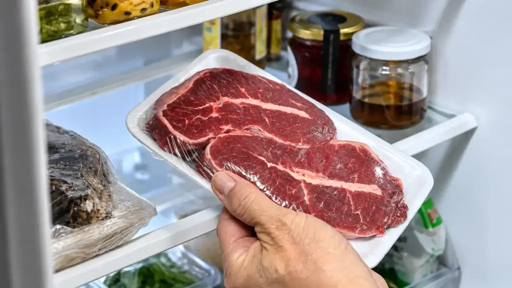 Una mano sostiene un paquete de bistec crudo —con la carne soltando líquido visible dentro del envoltorio— frente a un refrigerador abierto lleno de tarros, recipientes y diversos productos alimenticios.