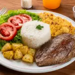 Día Mundial de la Alimentación: plato con arroz blanco, bistec, ensalada de hojas verdes y tomate, papas condimentadas y guarniciones sobre una mesa de madera