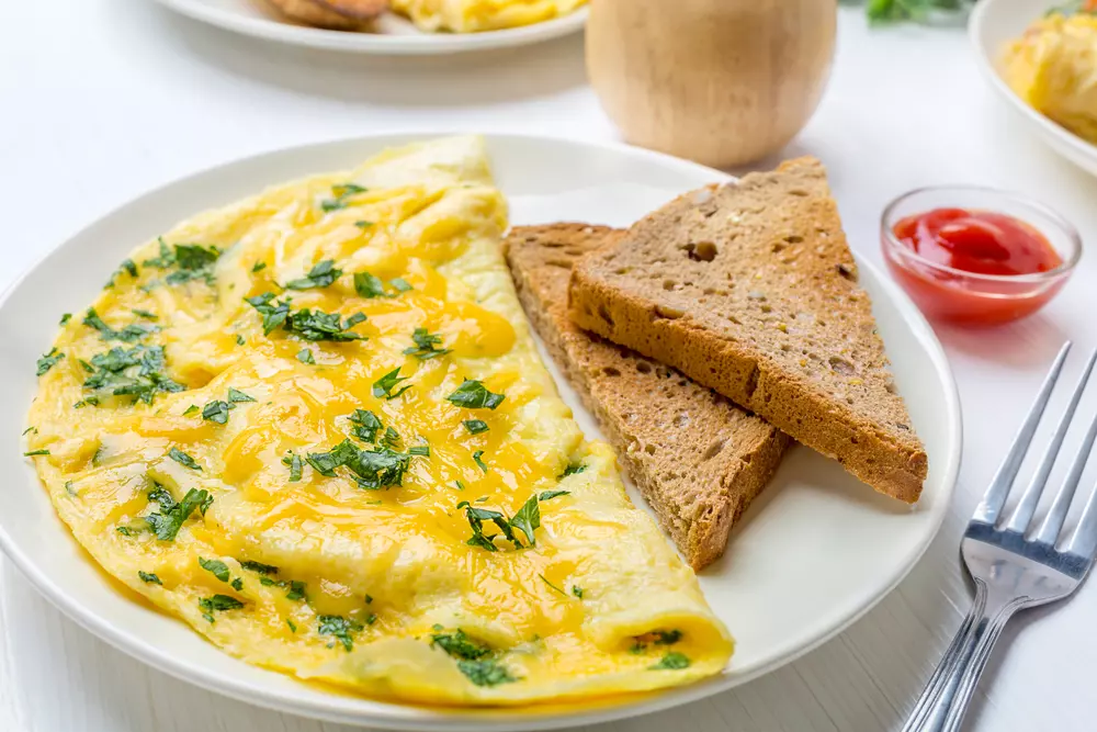 Uma bela omelete com queijo e salsinha salpicada por cima ao lado de duas torradas em formato de triângulo. A refeição está dentro de um prato branco em cima da mesa e ao lado um pequeno pote de molho vermelho para acompanhar.
