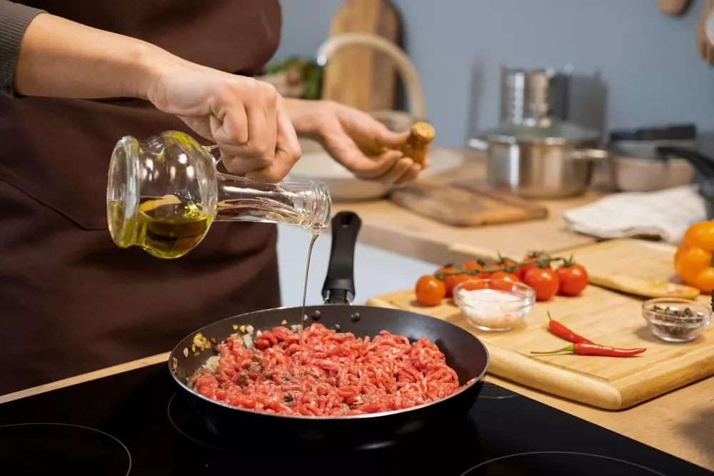 Mãos de uma mulher em sua cozinha preparando carne moída em uma frigideira e ao lado ingredientes para temperar e dar sabor ao refogado, como tomates, sal e pimenta vermelha.
