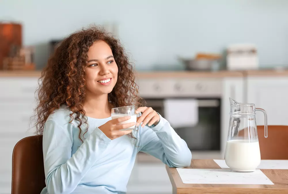 Uma mulher morena e de cabelos cacheados sentada na mesa de jantar de sua cozinha tomando um copo de leite com uma jarra do alimento também em cima da mesa. 