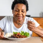 Uma senhora idosa e negra com expressão feliz almoçando um prato com carne moída, arroz, feijão e salada de alface e tomate. Trazendo o conceito de importância da carne vermelha em sua idade.