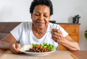 Uma senhora idosa e negra com expressão feliz almoçando um prato com carne moída, arroz, feijão e salada de alface e tomate. Trazendo o conceito de importância da carne vermelha em sua idade.
