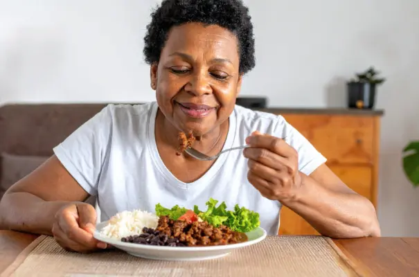 Uma senhora idosa e negra com expressão feliz almoçando um prato com carne moída, arroz, feijão e salada de alface e tomate. Trazendo o conceito de importância da carne vermelha em sua idade.