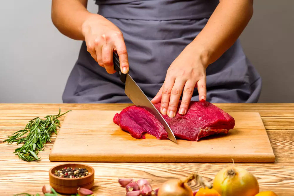 Pessoa cortando carne vermelha em uma tábua de madeira com utensílio de cozinha