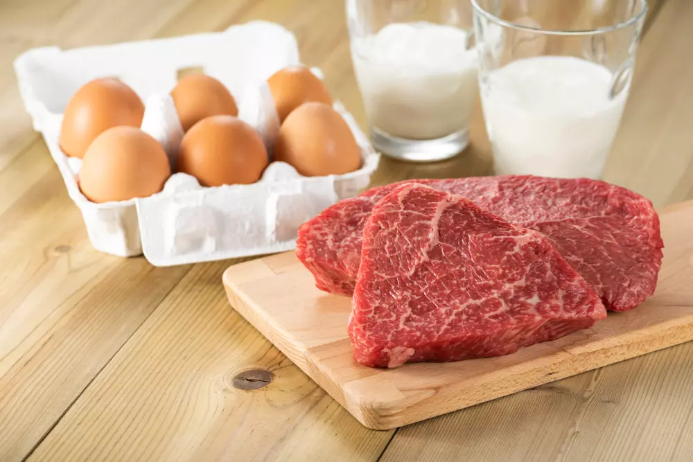 Em uma bancada de madeira clara está um cartela branca com ovos de galinha vermelhos, dois copos de leite de vaca e bifes  crus de carne vermelha.