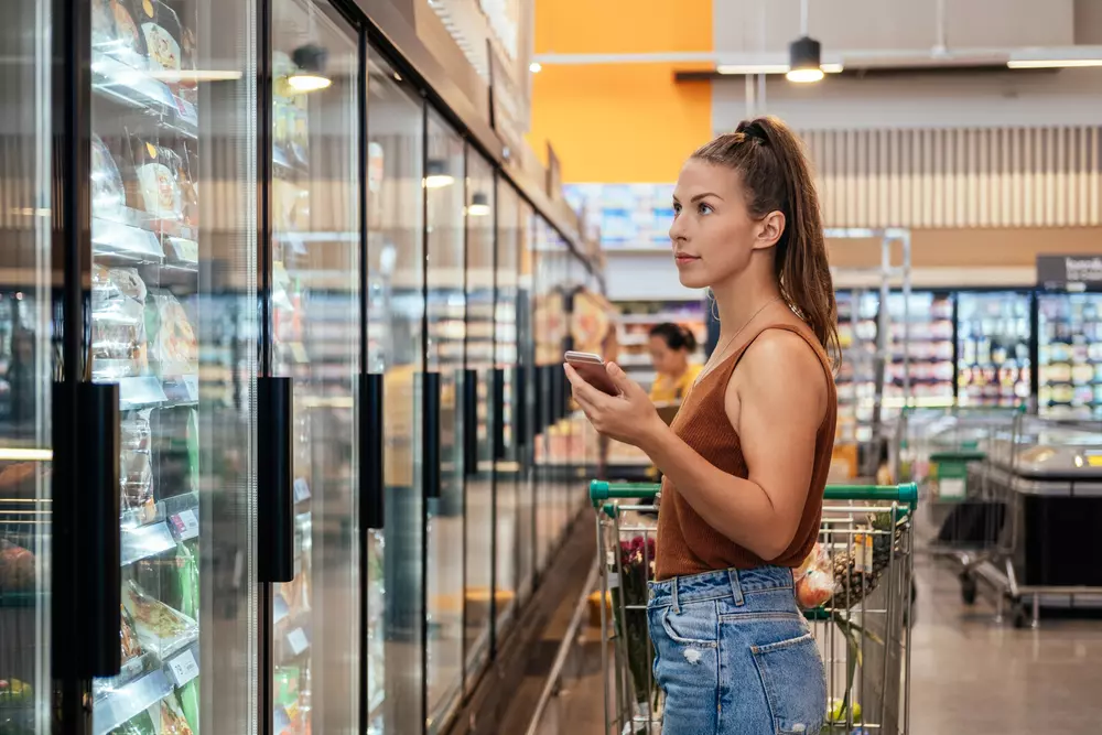 Mulher jovem fazendo compra em supermercado e usando smartphone ao lado de geladeiras de produtos embalados, interfaces com o consumidor.