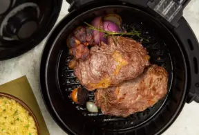 Uma peça de carne bovina dentro de uma air fryer sendo grelhada junto com cebolas e temperos.