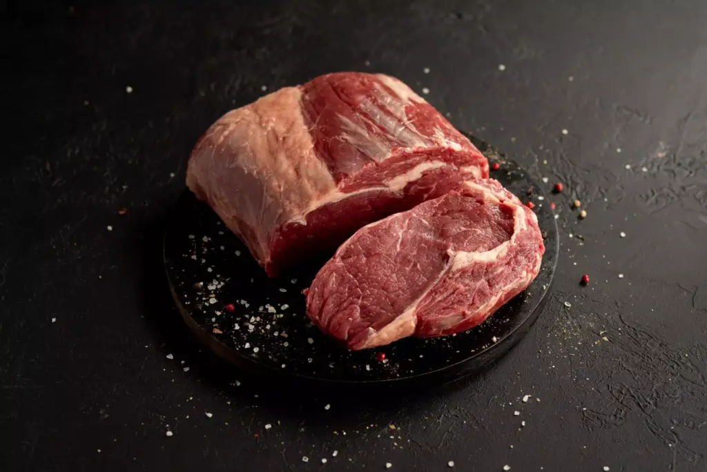 Carne bovina crua em cima de uma tábua escura e ao redor um pouco de sal e pimenta rosa.