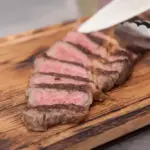 Bife de carne malpassado sendo cortado em uma tábua de madeira com utensílio de cozinha ao lado.