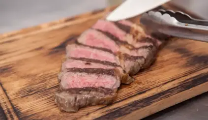 Bife de carne malpassado sendo cortado em uma tábua de madeira com utensílio de cozinha ao lado.