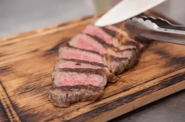 Bife de carne malpassado sendo cortado em uma tábua de madeira com utensílio de cozinha ao lado.
