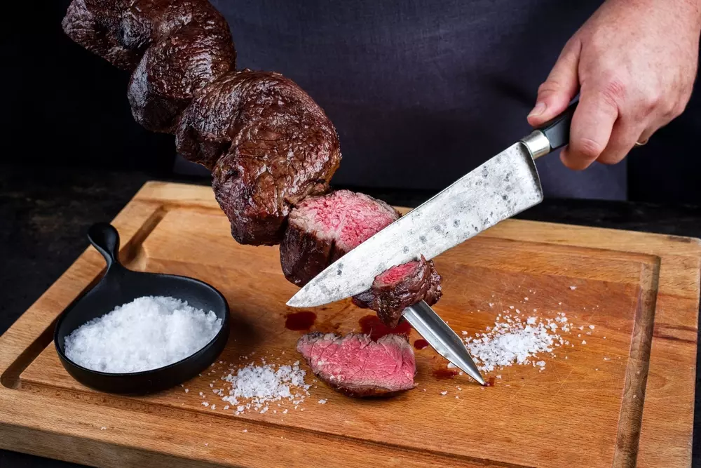 Person slicing a medium-cooked filet mignon on a wooden board, with salt to the left, prepared to serve a sophisticated meat dish.
