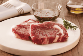 Bife Ancho in natura em corte amplo sobre uma tábua de madeira, acompanhado de molho e ramos de alecrim