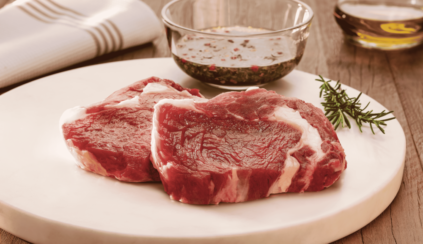 Bife Ancho in natura em corte amplo sobre uma tábua de madeira, acompanhado de molho e ramos de alecrim