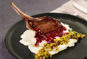 Imagem de carne de cordeiro assada acompanhada de purê de batatas, molho de frutas vermelhas e pistache, servido em prato preto para destaque da comida.