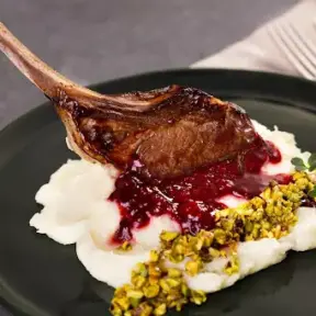 Imagem de carne de cordeiro assada acompanhada de purê de batatas, molho de frutas vermelhas e pistache, servido em prato preto para destaque da comida.