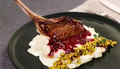 Imagem de carne de cordeiro assada acompanhada de purê de batatas, molho de frutas vermelhas e pistache, servido em prato preto para destaque da comida.