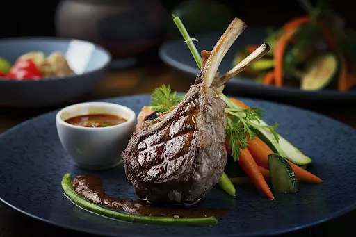 Carne de cordeiro assada, servida com molho e acompanhamentos de legumes frescos ao lado, em um prato preto elegante.