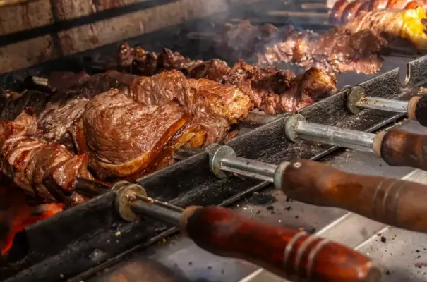 Churrasco sendo assado em uma churrasqueira, com diferentes tipos de carnes em espetos de metal e madeira, ideal para quem aprecia churrasco.